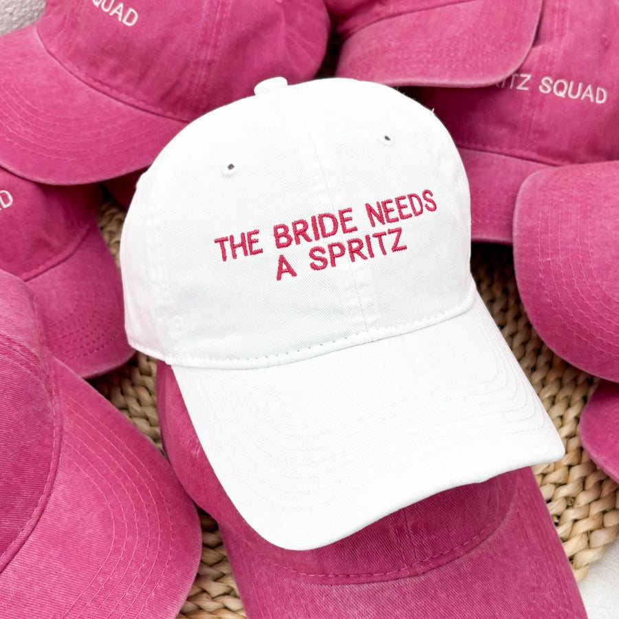 Casquette blanche avec le texte The bride needs a spritz parmi des casquettes roses