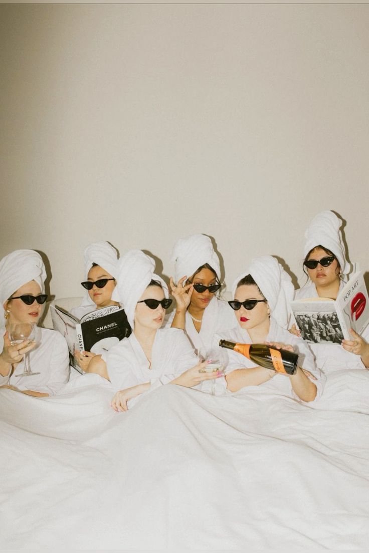 Amies en peignoirs blancs et lunettes de soleil sur un lit, avec du champagne