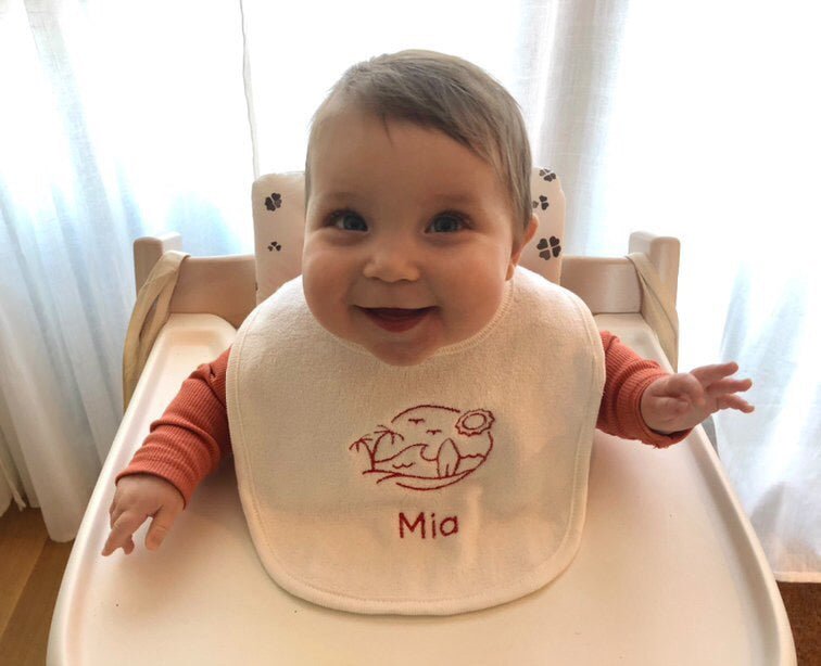 Bavetje - Tice Nits - Baby draagt een wit bavetje met rode tekst. Het slabbetje is gepersonaliseerd met een geborduurde afbeelding. Dit is het geboortekaartje van baby Mia